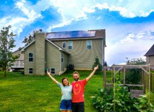 A couple stands in the backyard of their home making a smile shape with their arms. A solar array can be seen on the roof in the background.